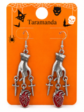A pair of gothic Halloween dangling earrings displayed on an orange backing card. The earrings feature silver-toned metal hands holding small silver crosses and red heart charms with white vein details.