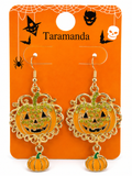 A pair of Halloween-themed dangling earrings displayed on an orange backing card. The earrings feature glittery orange jack-o'-lantern faces with gold filigree borders and small dangling pumpkins at the bottom.