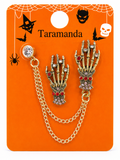A pair of skeleton hand brooches with gold-tone chains, red rhinestones, and clear crystal accents, displayed on an orange Halloween-themed backing card labeled 'Taramanda'.