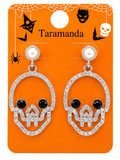 A pair of Halloween skull drop earrings on an orange display card. The earrings feature silver rhinestone-encrusted skulls with black gemstone eyes and nose, and white pearl studs.