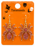 A pair of gold spider drop earrings covered in pink rhinestones, displayed on an orange Halloween-themed card labeled 'Taramanda'.