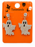 A pair of ghost-shaped dangle earrings covered in clear rhinestones with black eyes and mouths, hanging from gold-tone posts on an orange card printed with Halloween graphics and the brand name Taramanda.
