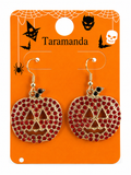 A pair of Halloween-themed dangle earrings shaped like jack-o'-lanterns, covered in red rhinestones with gold-tone metal outlines and hooks. They are displayed on an orange card printed with spiderwebs, black witch silhouettes, and the brand name 'Taramanda'.