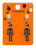 A pair of Halloween skeleton drop earrings displayed on an orange Taramanda card. The earrings feature black skeleton charms wearing red Santa hats and green bow ties, with rhinestone-studded round studs.