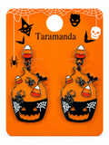 A pair of Halloween-themed dangling earrings displayed on an orange backing card. The earrings feature black jack-o'-lantern faces with white spiderweb details, candy corn accents, and small black spiders.