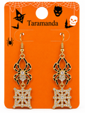 A pair of gold-tone spider drop earrings with black spider body details and rhinestone-embellished web motifs, displayed on an orange Halloween-themed card labeled Taramanda.