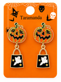 A pair of Halloween-themed dangle earrings on an orange display card. The earrings feature round orange pumpkin tops with black jack-o'-lantern faces, outlined with gold and green rhinestone-like detailing, and drop down to black shopping-bag charms with white ghosts and gold stars.