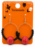 Halloween hoop earrings with black beads and red spiderweb-patterned beads, displayed on an orange Taramanda brand card with spider-themed graphics.