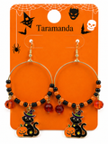 A pair of Halloween hoop earrings on an orange retail display card. The earrings have gold-tone hoops with black and orange beaded strands, faceted orange and red beads, and dangling black cat charms wearing witch hats with pumpkin accents.