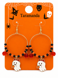 Halloween hoop earrings with gold-tone metal hoops, black and orange beads, red faceted beads, and white ghost charms holding pumpkins, displayed on an orange Taramanda backing card.