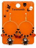 A pair of Halloween hoop earrings on an orange retail display card. The earrings have gold-tone hoops, orange and black beads, red faceted beads, and black bat-shaped charms with white skull details.