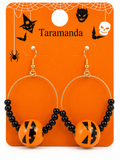 A pair of Halloween hoop earrings on an orange retail display card. The earrings have gold-tone hoops, black beads, and round orange beads printed with a black jack-o'-lantern face.