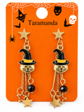 A pair of gold-toned dangle earrings displayed on an orange backing card. Each earring features a cat face wearing a black witch hat with a yellow band, with hanging star charms and black beads.