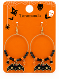 A pair of Halloween hoop earrings displayed on an orange backing card. The earrings have gold-tone hoops, black and orange beads, and black bat charms with gold details.