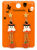 A pair of Taramanda brand Halloween dangle earrings displayed on an orange backing card. The earrings feature gold-plated chains and star studs, a white ghost wearing a black witch hat with orange trim, and a dangling orange pumpkin charm with a green stem.