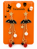 A pair of Halloween-themed bat drop earrings on an orange display card. The earrings feature gold-tone metal chains, black enamel bat wings with small gold stars, gold star charms, white pearl beads, and red faceted beads at the bottom.