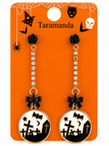 A pair of Halloween-themed dangle earrings on a branded orange backing card. The earrings have black rose studs, gold-tone chains with clear rhinestones, black bow charms, and round white-and-black tombstone pendants featuring bats, crosses, and a crescent moon.