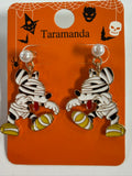 A pair of Mickey Mouse mummy earrings on an orange display card. The earrings feature a mummy with black and white striped wrap, a red tongue sticking out, and yellow feet, hanging from pearl-like studs.