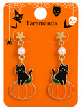 A pair of Halloween-themed dangle earrings displayed on an orange backing card. The earrings feature black cat and pumpkin engravings with gold-tone metal, a white faux pearl, and a glittery gold star.