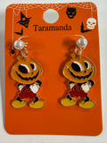 A pair of Halloween-themed earrings feature a jack-o'-lantern head with black eyes, a red shorts outfit, and yellow shoes, suspended from white faux pearls.