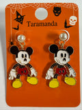 A pair of Halloween earrings on an orange Taramanda display card. The earrings feature red shorts, yellow shoes, and black-and-white ears, with pearl-like white studs at the top.