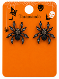A pair of black spider-shaped stud earrings with faceted dark gray rhinestones, displayed on an orange Halloween-themed card labeled 'Taramanda' with spider, bat, witch hat, skull, and spiderweb graphics.