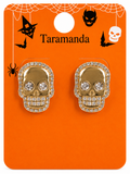 A pair of gold skull stud earrings encrusted with clear rhinestones, displayed on an orange Halloween-themed backing card with the brand name 'Taramanda'.
