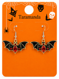 A pair of bat-shaped drop earrings displayed on an orange Halloween-themed card. The earrings have black wings, gold outlines, and red gemstone accents.