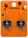 A pair of gothic Halloween dangle earrings featuring black and silver skull and cat heart charms on silver chains, displayed on an orange backing card with spiderwebs, bats, and the brand name 'Taramanda'.