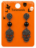 A pair of gothic Halloween dangle earrings on an orange display card. The earrings feature round studs with black crystal pavé skulls, a single black bead, and a larger black crystal pavé skull dangle.