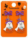 A pair of Halloween-themed dangle earrings on a branded orange backing card. Each earring has a purple bow and a ghost-shaped charm wearing a purple witch hat and holding a glowing orange cauldron with blue steam.
