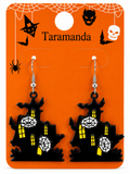 A pair of Halloween dangling earrings shaped like haunted houses, displayed on an orange card with spider, bat, witch, skull, and bat graphics. The card is labeled 'Taramanda'.