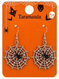 A pair of silver spiderweb dangle earrings with black spider centers, displayed on an orange Halloween-themed backing card labeled 'Taramanda'.