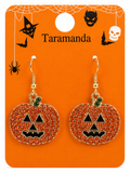 A pair of Halloween pumpkin drop earrings with gold-tone hooks and rhinestone-embellished orange jack-o'-lantern faces, displayed on an orange Taramanda retail card with spiderwebs, bats, and skull graphics.