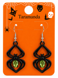 A pair of black spider-shaped drop earrings with green teardrop gemstones, displayed on an orange Halloween-themed card labeled 'Taramanda'.