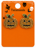 A pair of glittery gold jack-o'-lantern pumpkin stud earrings with green glittery stems and black enamel facial details, displayed on an orange Halloween-themed backing card labeled 'Taramanda'.