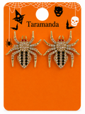 A pair of spider-shaped earrings with gold-tone metal bodies, clear rhinestones, and black enamel stripes, mounted on an orange Halloween-themed display card labeled Taramanda.
