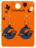 A pair of Halloween-themed dangling earrings on an orange display card. The earrings feature dark blue crescent moon pendants with purple bat wings and black bats, attached to silver hooks.