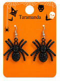 A pair of black spider drop earrings with rhinestone-embellished bodies and silver-tone hooks, displayed on an orange Halloween-themed card labeled Taramanda.